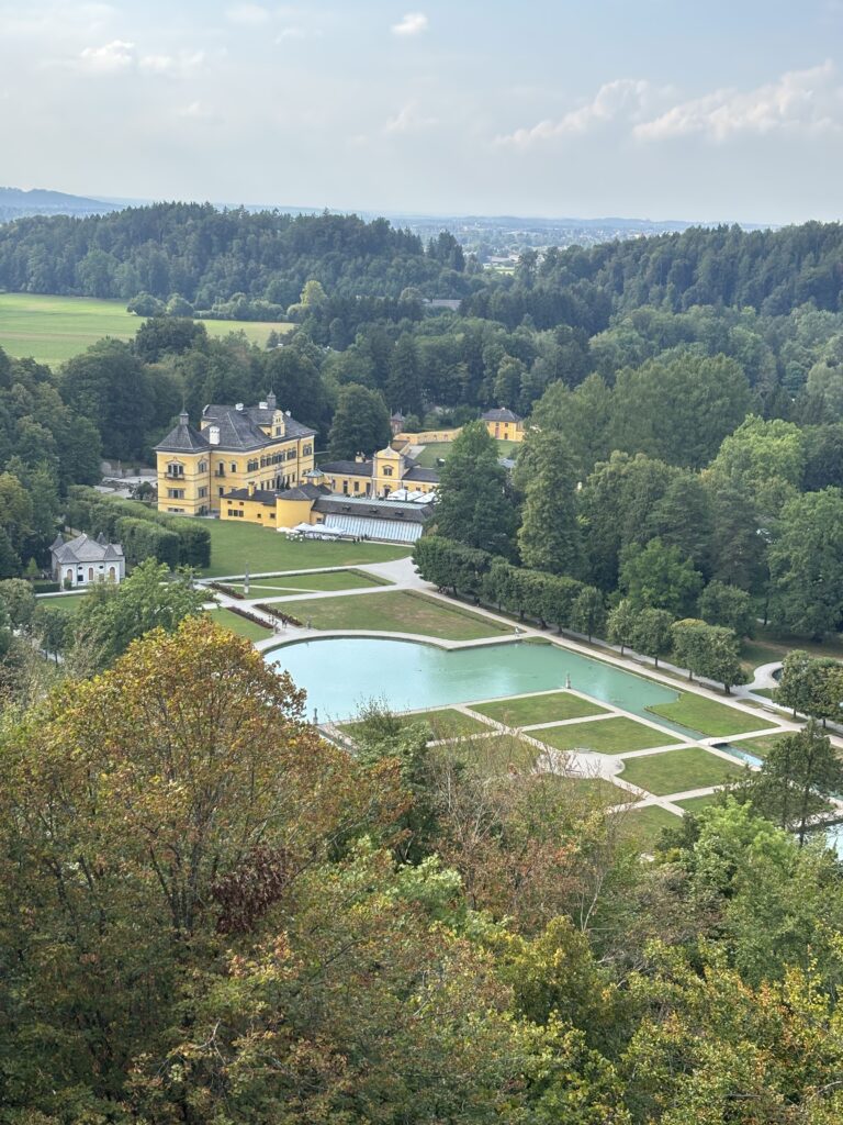 Over Head View of Hellbrunn Palace and Gardens taken from the nearby Woodland Viewpoint. This includes a bright yellow palace and manicured gardens with a light blue pond.