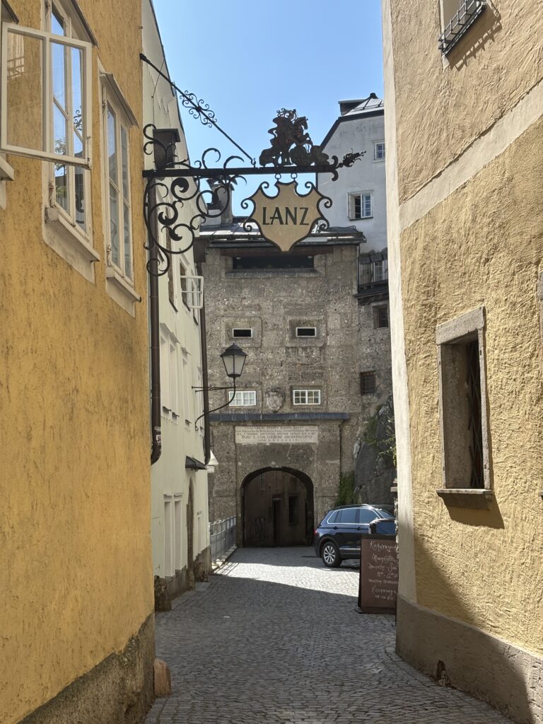 A medieval street with cobbled paths, colourful old stone houses, an old metal sign featuring the word 'Lanz'. The street leads to an old bridge tower.