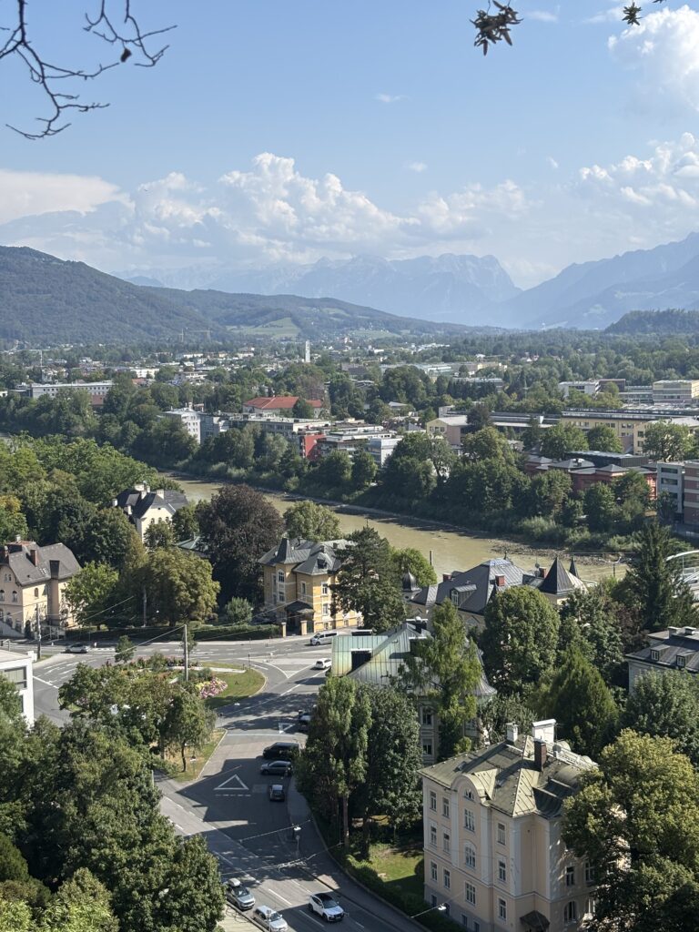 A panoramic bird's eye view down to Salzburg Old Town, Salzach River, and the mountain ranges of Gaisberg and Untersberg. It is a sunny, clear day and the river is a vibrant green hue.