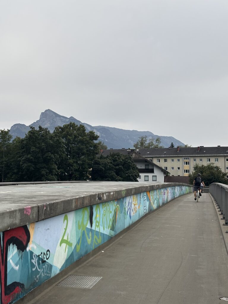 A bridge with colourful graffiti featuring a cyclist crossing the Salzach river. The Untersberg mountain can be seen in the distance.