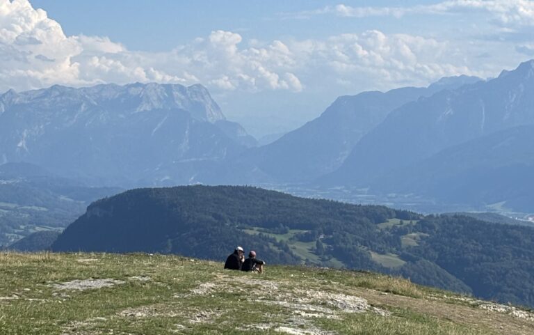 Two people sitting and taking in the panoramic mountain views from Gaisberg Peak overlooking the neighbouring Untersberg Mountain range and surroundings area of Salzburg.